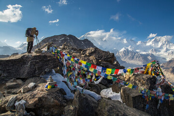 A photographer with a backpack stands on top of a mountain and shots the mountains around. Prayer flags are on the stones. Snow-capped peaks are on the background. Blue sky with a few clouds.