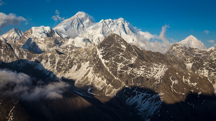 Berge Achttausender der Everest-Region vor Sonnenuntergang. Everest und Lhotse in der Mitte, Makalu rechts. Strahlend blauer Himmel oben mit leichten Wolken. Unten dunkle Schatten.
