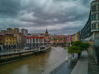 Cityscape of Bilbao, Spain, with the market hall and the spire of the church San Anton aside the river Nervion with colorful buildings and a dark cloudy sky