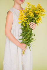 Obraz na płótnie Canvas Girl in white linen dress posing on a green background with a bouquet of yellow wildflowers