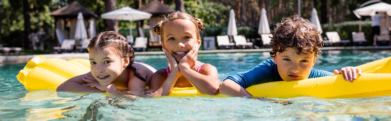 horizontal image of joyful friends looking at camera while floating on inflatable mattress in pool