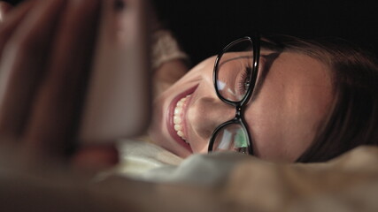 Woman with glasses looks at smart phone and lies on bed, smile on face