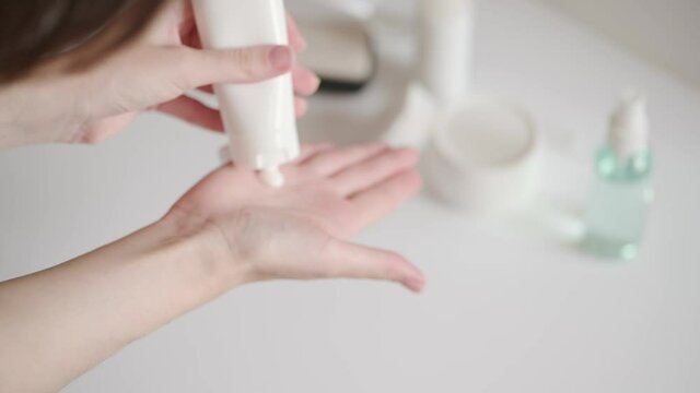 The woman takes a plastic tube of cream from the white table and smears her hands. Hand skin care at home. Moisturizing lotion with vitamins, body care. Cosmetic beauty skincare product.