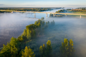 Fototapeta na wymiar Early morning landscape. Foggy river. Highway bridge across the Volkhov river. River valley in the morning fog at sunrise. View from above. Rays of the sun breaking through the fog in over the trees