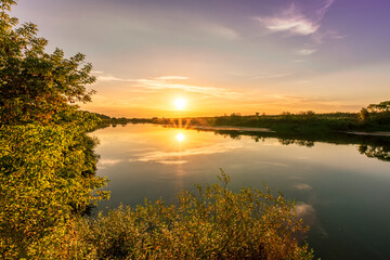 Obraz na płótnie Canvas Scenic view at beautiful summer river sunset with reflection on water with green bushes, grass, golden sun rays, calm water ,deep blue cloudy sky and glow on a background, spring evening landscape