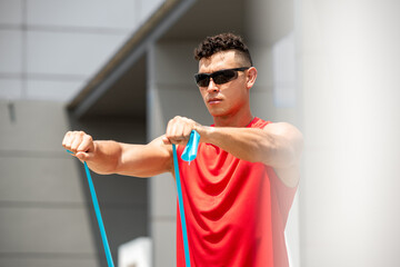 Handsome sports man doing shoulder front raise exercise with resistance band outdoors in the sun