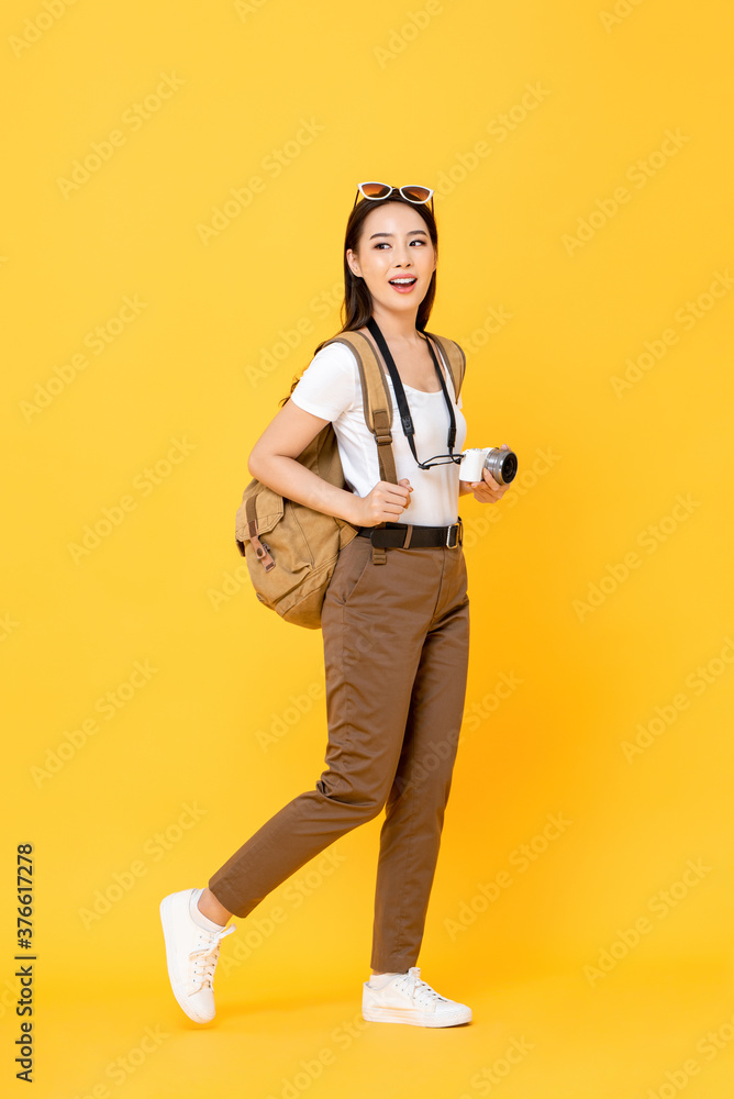 Wall mural full length portrait of smiling cute young asian woman tourist with camera waling on isolated yellow