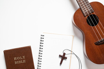 Top view of ukulele, Holy Bible, and notebook with wooden cross on white background