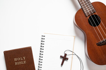 Fototapeta premium Top view of ukulele, Holy Bible, and notebook with wooden cross on white background