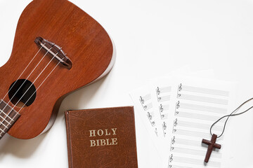 Top view of ukulele with blank stave pad bar for music notes, Holy Bible, and wooden cross on white background