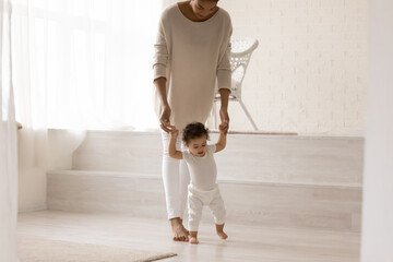 Caring young african american mum holding hands of little curly cute biracial toddler boy or girl, teaching making first steps in living room. Cute small son or daughter walking with mother's help.