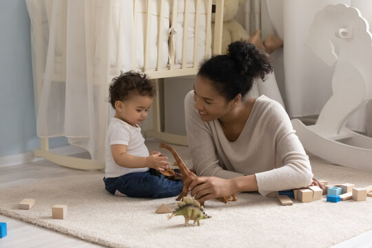 Caring young happy african ethnicity mother or babysitter relaxing in children bedroom with cute curious small biracial boy or girl, playing toys together on carpet, developing imagination creativity.