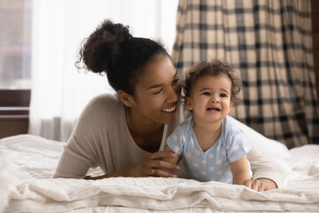 Head shot affectionate loving young mixed race woman lying on bed with cute baby, comforting for daytime sleeping. Caring biracial mommy cuddling small adorable kid son or daughter in bedroom.