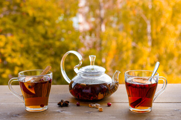 Autumn tea party outdoors. Glass kettle with hot herbal tea and two glass cups on a wooden table against the background of yellow trees. 