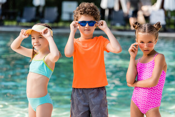 curly boy in t-shirt and girls in swimsuits touching sunglasses near pool