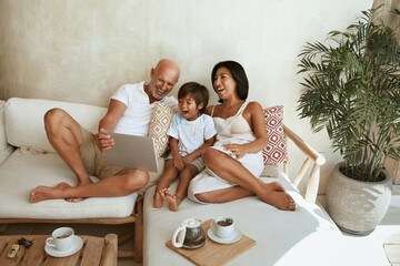 Happy Family On Sofa. Portrait Of Mixed Race Parents With Little Son Using Tablet And Enjoying Leisure On Summer Vacation At Tropical Resort. Different Ethnic Mom And Dad With Boy On Weekend.