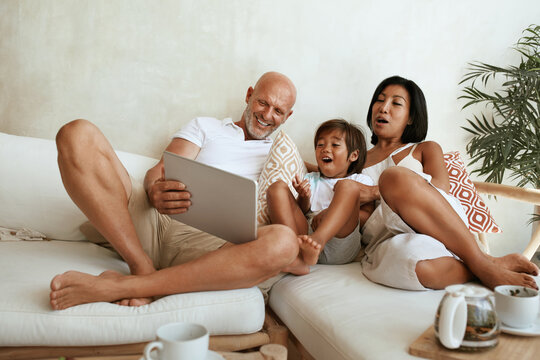 Family Using Tablet On Weekend. Happy Interracial Parents With Little Boy Sitting On Sofa, Using Portable Digital Device And Having Fun At Home. Mom, Dad And Son On Summer Vacation At Tropical Resort.