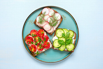 Variety of sandwiches for breakfast with slice of whole grain dark bread, cream cheese, cucumbers, radishes, cherry tomatoes, garnished with dill, green onions on light blue table Top view Flat lay