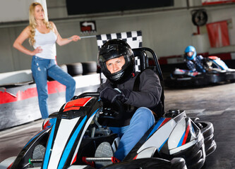 Diligent efficient positive man in helmet driving car for karting in sport club, friendly woman with flag on background