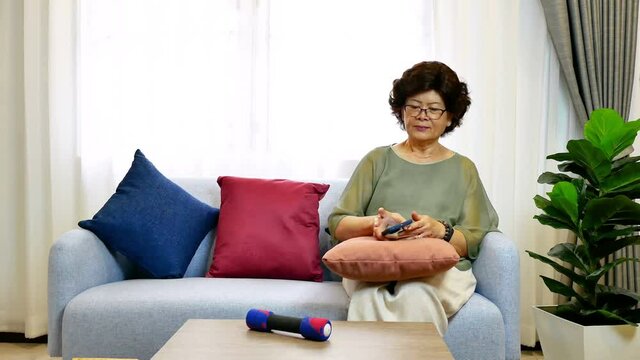 Asian senior Woman using mobile phone , sitting on sofe in living room, after that lifting little dumbbell for relaxation. Older people doing exercise at home concept.