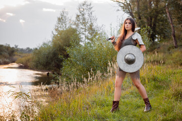 Medieval warrior woman with war paint, in chain mail armor with a sword and a shield in her hands, posing standing against the backdrop of bokeh sunset, river and forest. Armed fantasy girl. © Михаил Гута