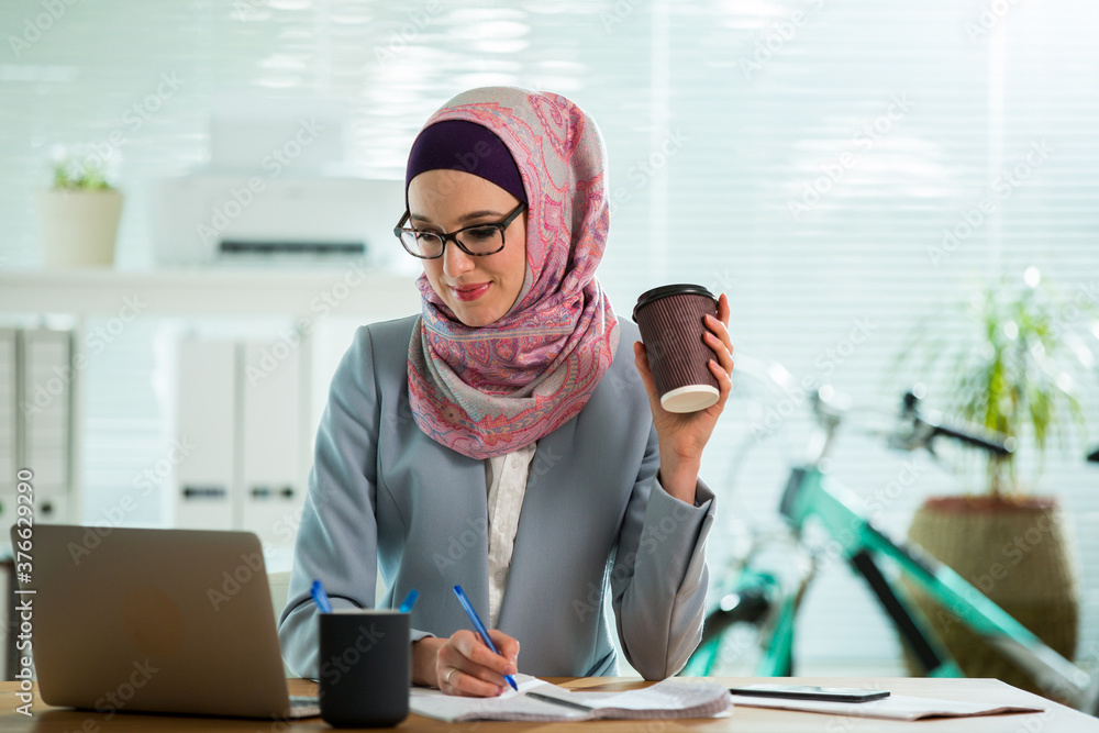 Wall mural beautiful young working woman in hijab, suit and eyeglasses sitting in office, smiling. portrait of 