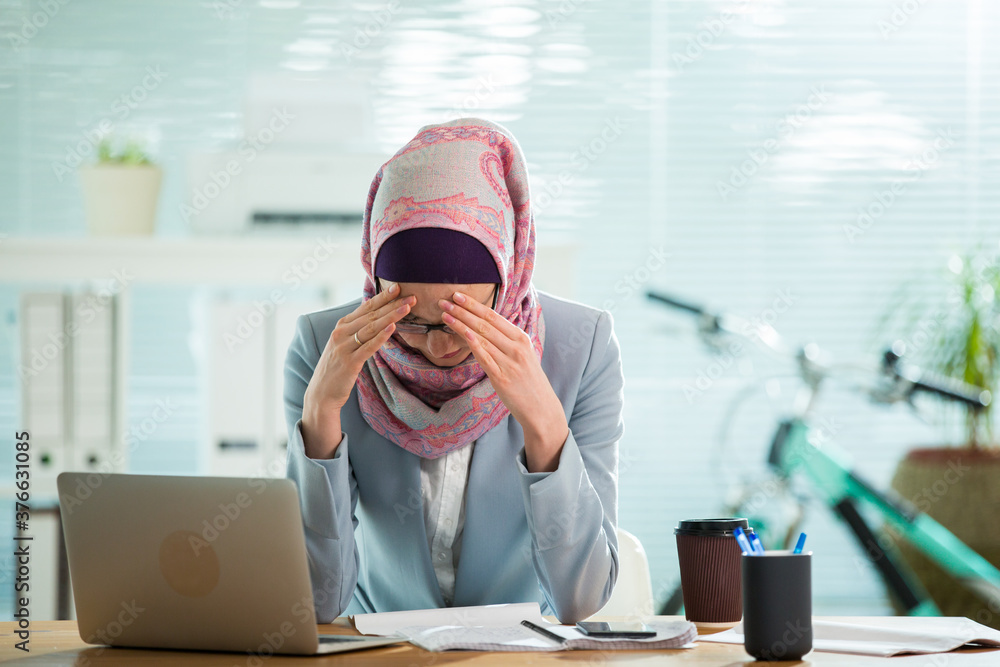 Wall mural beautiful young working woman in hijab, suit and eyeglasses sitting in office, having headache. mode