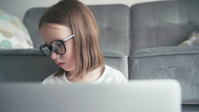 A close-up shot of a serious little 8 years old girl in glasses looking to a laptop screen and typing. Remote education technologies and homework.