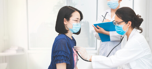 Female doctor use stethoscope do medical checkup to woman in hospital, trainee stand beside taking note on information chart. wearing medical mask to protect from spreading virus, healthcare concept