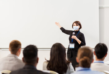 business, education and pandemic concept - smiling businesswoman or teacher in mask with microphone talking to group of students at conference, presentation or lecture and showing something on screen