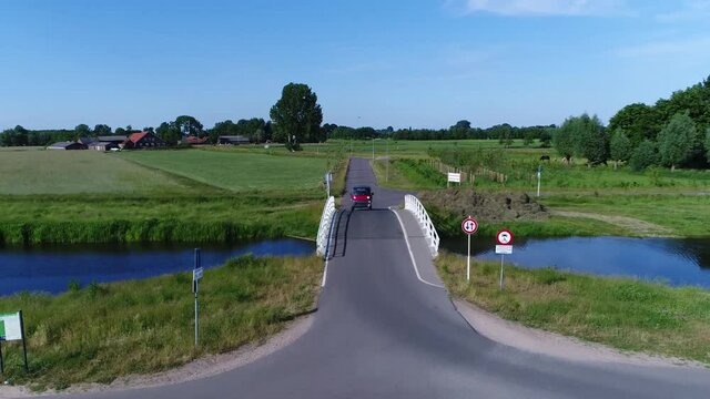 Low altitude aerial view of red microcar driving over small bridge drone moving above road and vehicle scenery polder landscape also showing farm in the background 4k high resolution footage