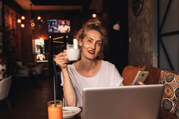 Portrait of smiling senior female blogger in casual wear with laptop and smartphone sitting in cafe and looking at camera.