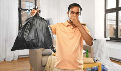 recycling, sorting and sustainability concept - young indian man holding stinky trash bag over messy home room background