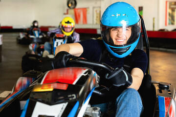 Glad smiling girl and her friends competing on racing cars at kart circuit - obrazy, fototapety, plakaty