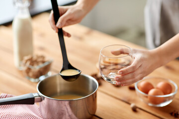christmas and seasonal drinks concept - close up of hands with ladle pouring eggnog from pot to glass at home