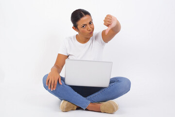 Discontent European woman shows disapproval sign, keeps thumb down, expresses dislike, frowns face in discontent, dressed in white shirt, isolated over gray background. Body language concept.