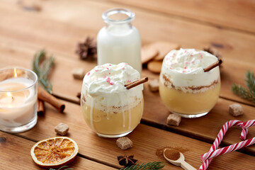 christmas and seasonal drinks concept - glasses of eggnog with whipped cream topping and cinnamon, fir tree branches, brown sugar and candle burning on wooden background