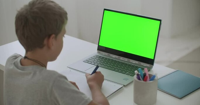 little boy is drawing sitting at table with laptop with green screen for chroma key technology, child at home at weekend