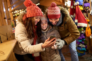 Fototapeta premium family, winter holidays and technology concept - happy mother, father and little daughter with smartphone at christmas market in evening