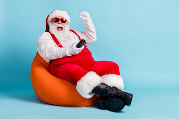 Full length photo of retired old man grey beard joystick crazy win playstation against boss sit beanbag wear red santa x-mas costume suspender sunglass cap isolated blue color background