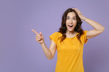 Surprised young brunette woman 20s wearing basic yellow t-shirt posing pointing index finger aside on mock up copy space put hand on head isolated on pastel violet colour background, studio portrait.