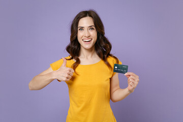 Smiling funny young brunette woman 20s wearing basic yellow t-shirt posing hold in hands credit bank card showing thumb up looking camera isolated on pastel violet colour background, studio portrait.