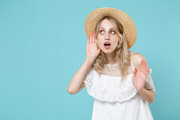 Shocked curious young blonde woman 20s wearing white summer dress hat standing try to hear you overhear listening intently looking aside isolated on blue turquoise colour background studio portrait.