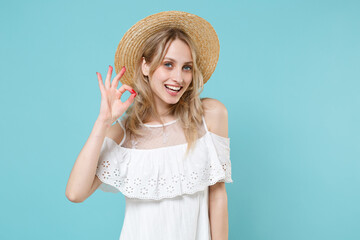 Smiling charming beautiful attractive young blonde woman 20s wearing white summer dress hat standing showing OK gesture looking camera isolated on blue turquoise colour background, studio portrait.