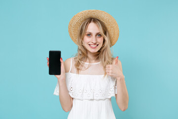 Smiling young blonde woman 20s in white summer dress hat hold mobile phone with blank empty screen mock up copy space showing thumb up isolated on blue turquoise colour background studio portrait.