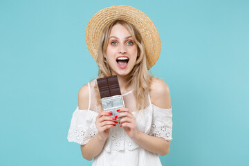 Shocked excited young blonde woman 20s wearing white summer dress hat standing holding chocolate bar keeping mouth open looking camera isolated on blue turquoise colour background studio portrait.