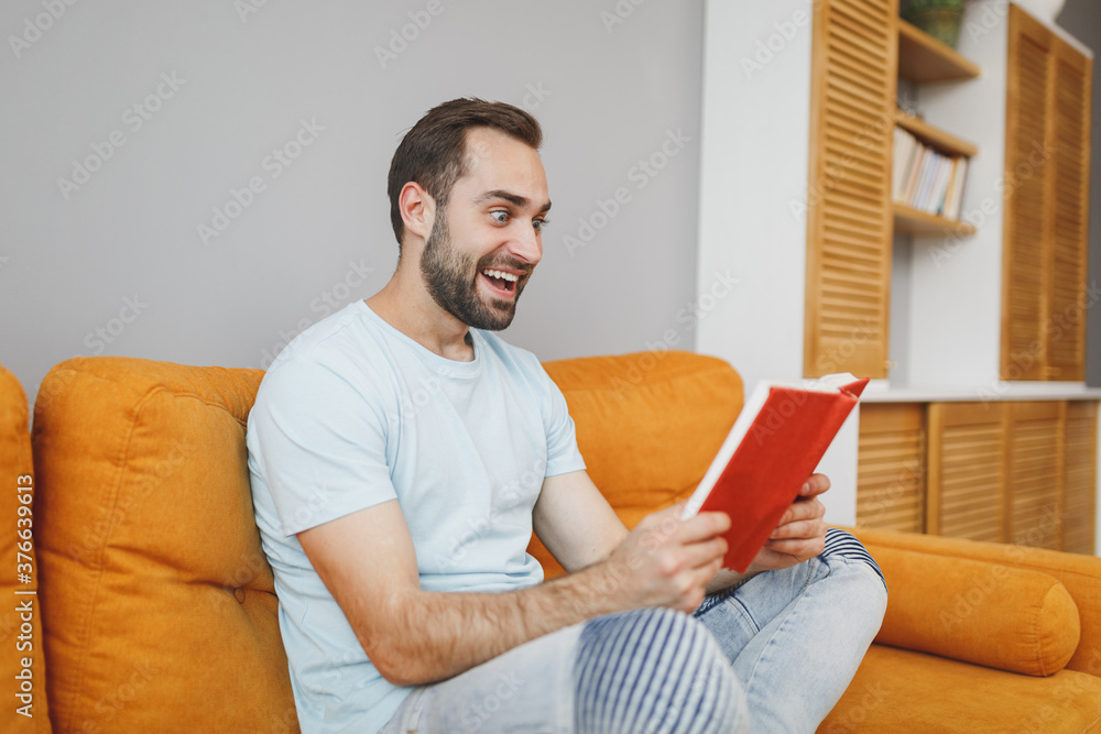 Wall mural Excited cheerful funny attractive young bearded man 20s wearing casual blue t-shirt holding in hands reading book looking aside sitting on couch resting relaxing spending time in living room at home.