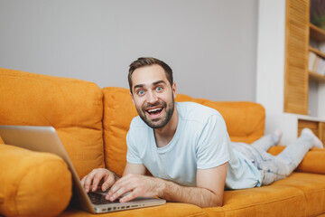 Shocked amazed surprised attractive young bearded man 20s wearing casual blue t-shirt working on laptop pc computer lying on couch resting relaxing spending time in living room at home.