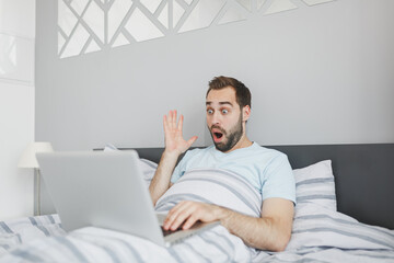 Shocked young bearded man 20s in basic blue t-shirt working on laptop pc computer spreading hands lying in bed with striped sheet pillow blanket resting relaxing spending time in bedroom at home.