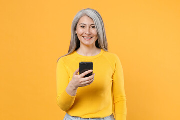 Smiling pretty beautiful gray-haired asian woman wearing casual clothes standing using mobile cell phone typing sms message looking camera isolated on bright yellow colour background, studio portrait.